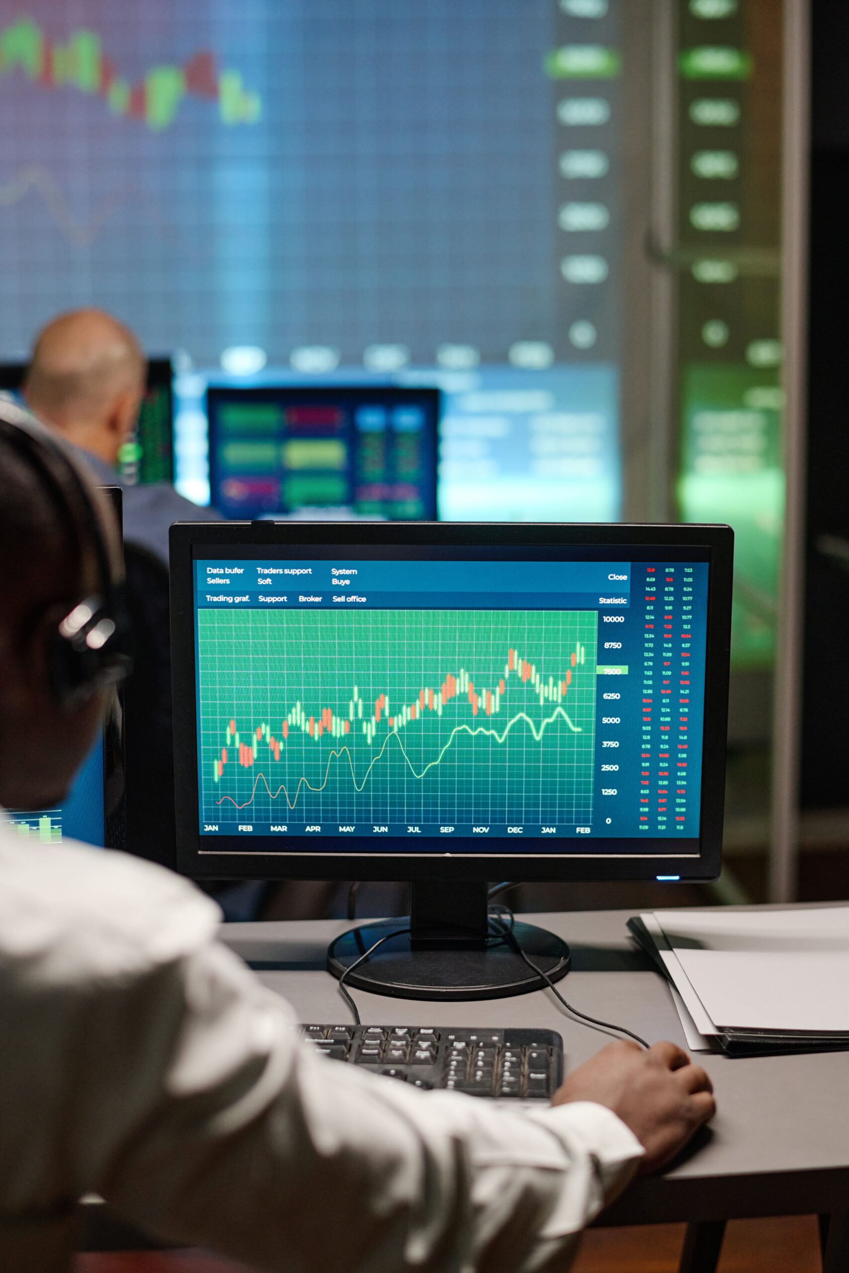 Man Working With Stock Trading Data Rear view of unrecognizable African American man sitting in front of computer monitors watching stock trading charts in evening